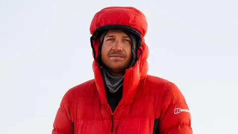 Berghaus Former Rugby player and explorer Ed Jackson stands in Nepal wearing a red snowproof puffa jacket. 