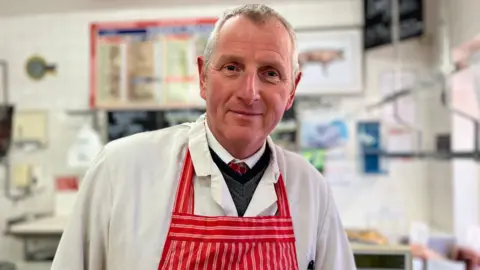 A man with grey hair is wearing a white jacket and a red striped apron.
There are information notices on a white wall behind him.