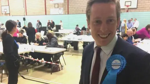 BBC Tom Pursglove with short fair hair wearing a blue jacket and red tie with a blue rosette smiling at an election count