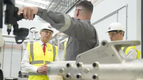 PA Media A man in a hard hat and safety jacket looks thoughtfully as another man in a grey boiler suit points at a machine