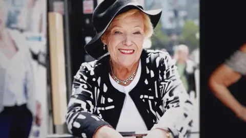 Old woman with blonde hair wearing a black hat and a black and white jacket 