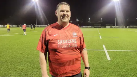 Coach Richard Perry wearing a Saracens Foundation branded rugby training shirt and standing in front of the pitches at North Bristol Rugby Club.