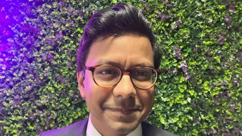 Narayan wearing a grey suit and round glasses. He has black hair.