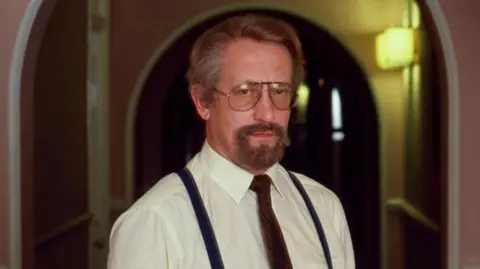 Getty Images Oleg Gordievsky looks at the camera, he is wearing glasses and has a beard and a moustache. He is wearing a white shirt, black braces and a brown tie and the background behind him is an arched corridor painted in a pale pink.