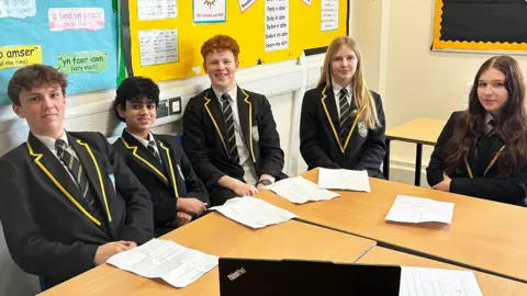 Five Year 10 students sit around two desks pushed together. They all wear a white shirt, a navy, white and gold striped tie, navy blazer with gold edging on the collar and a school badge on their left. Kit is white with short light brown hair. Jake has olive skin with short black hair. Hugo is whit with slightly curly red hair which is cropped at the sides. Cali is white with long blonde hair. Maisy is white with very long brown hair which is slightly wavy at the bottom. There are pinboards with phrases in Welsh behind them.