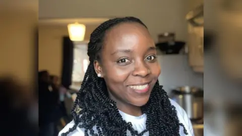 A woman smiles at the camera as she stands inside her home. She is wearing a white top and pearl earrings.