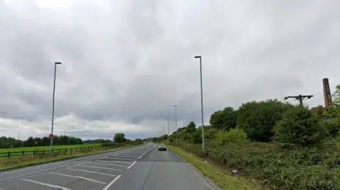A tarmac road sweeps between a green field on one side and a large hedge on the other. 