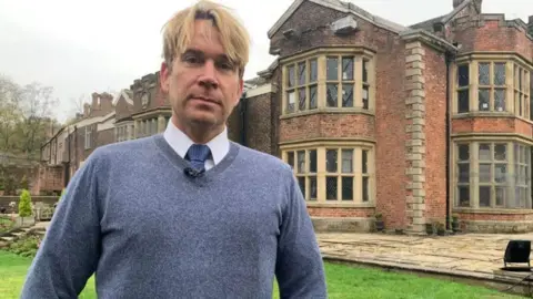 The US actor Hopwood DePree outside Hopwood Hall in Middleton. He is wearing a blue v-neck jumper, white shirt and blue tie. He has short-mid length blond hair 