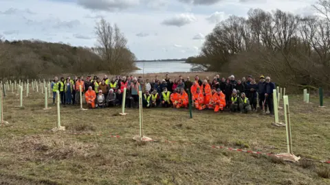 Creating Nature's Corridors A large group of people are standing together in front of a reservoir, surrounded by woodland. Some of them are wearing yellow and orange hi-vis jackets. There are several newly planted trees surrounding them.