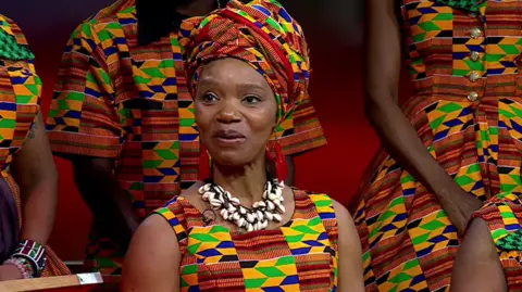 Anna Mudeka, wearing a dress and headdress with a yellow, green, blue and red pattern and a shell necklace, smiles towards a camera while sat down, with other members of a choir wearing similar clothing can be seen behind her. 