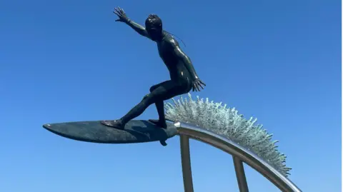 A bronze statue of a surfer wave riding. Its dark and taken on a bright sunny day with blue skies in Newquay.