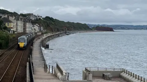 BBC Dawlish - houses, a train, a sea wall and the sea on a stormy day
