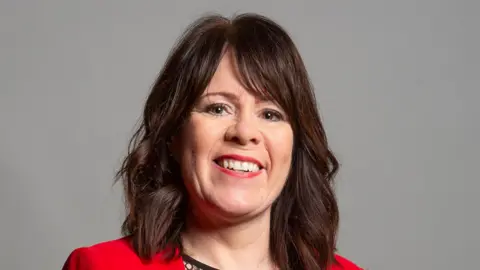 House of Commons Mary Foy has brunette hair and is looking directly at the camera smiling. She is wearing a red jacket and red lipstick. 