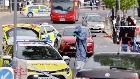 A forensic officer stands in a pale blue protective suit surrounded by police vehicles. A group of police officers can be seen in the background