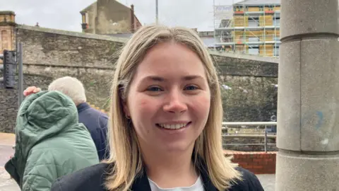 Shows a young woman with blond hair smiling and a man and woman in the background and behind them the Derry walls