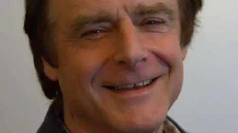 Isle of Wight Council Phil Jordan smiles at the camera, against a grey background. He has slightly greying brown hair and wears a dark top.