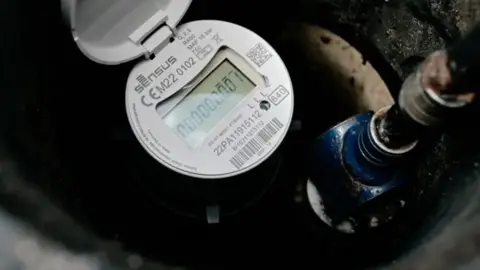 Essex and Suffolk Water We can see part of a smart meter. A white circle with numbers, branding and a barcode on it. In the middle, there's a digital display.