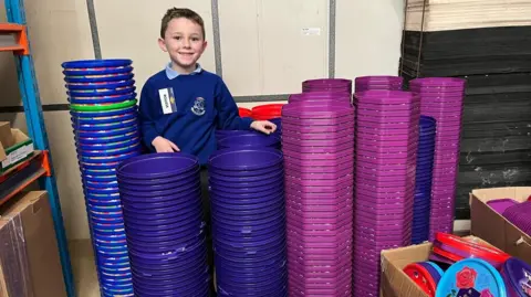 Teddy is in a blue school jumper. He has towers of sweet tubs around him. Their lids are in cardboard boxes. He may be in his home garage.