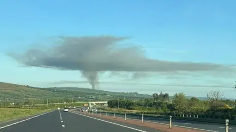 a large plume of smoke above fields. In the foreground is a dual carriageway