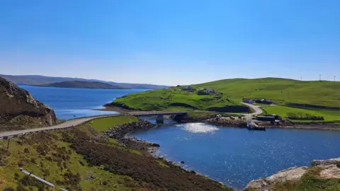 Sylvia Anderson The green Shetlands Islands with a bridge connecting two grassy regions. The water is sparkling blue and it's a clear blue day.