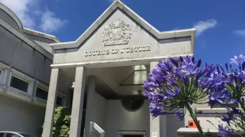 Outside Truro Crown Court. The building is light grey and has the words on the front 'Courts of Justice'. There are purple flowers on the right.
