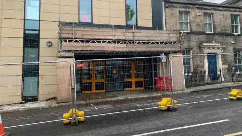 Durham Bus Station in Durham. The entrance has wooden scaffolding covering the building and looks like it is awaiting repairs. There is large metal fencing protecting the entrance.