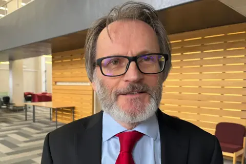 BBC/David Macmillan Stephen Harker has browny-grey hair, a grey beard, glasses and is wearing a black formal jacket, blue shirt and red tie. He is standing indoors. There is a table and a chair behind him.