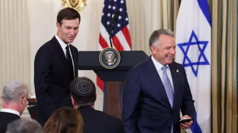 Reuters Jared Kushner, wearing a dark suit and tie, and Steve Witkoff, wearing a dark blue suit and light blue tie, walk during a news conference. There is an American and Israeli flag in the background.