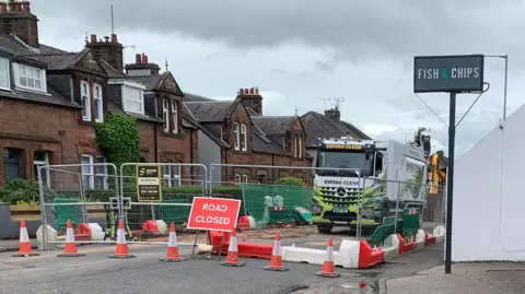 A road closure outside a chip shop - an area of the road has been fenced off with work taking place on the ground. There is a lorry within the work area.