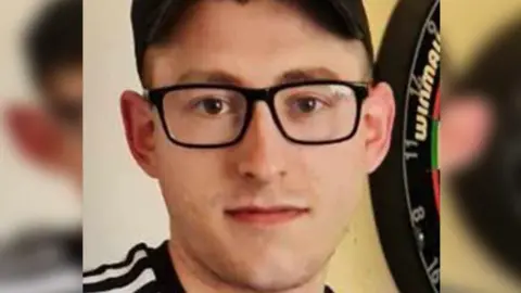 Essex Police Martyn Richardson, wearing glasses and a cap, looking at the camera. He is standing by a dart board and wearing a black and white top.