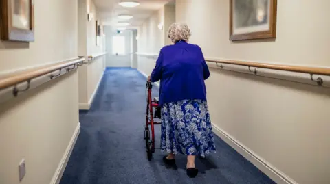 An elderly woman walks down a corridor in a care home with her back to the camera. She is using a walker for support and is wearing a blue cardigan and a long blue flowery skirt. The walls are cream and the carpet is blue.