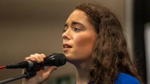 Handout Eimear is onstage singing. Her brown hair is wavy. She's holding a mic and is wearing blue. 