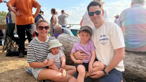 BBC/Kate Justice Meg is on the left and wearing a black and white hooped top and holding Charlie, who is wearing a light brown sun hat and striped top. Eddie is on the right and wearing a white T-shirt and dark glasses and holding three-year-old Henry, who has a baseball cap on and a purple t-shirt with black short.