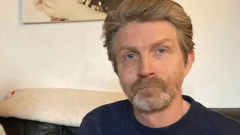 Anthony Mills pictured with swept back brown hair wearing a blue sweatshirt. he is sat on a black leather sofa.