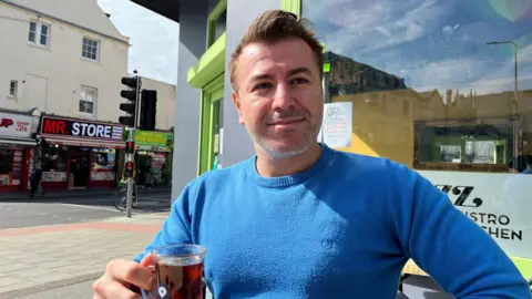 George Carden/BBC Emre Durmaz sits outside his café with Western Road behind him, wearing a blue jumper and holding a cup of tea.