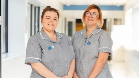Two women in grey nurses' scrubs stand together side by side and smile at the camera. They both have a blue dementia pin in the shape of a flower on their scrubs.