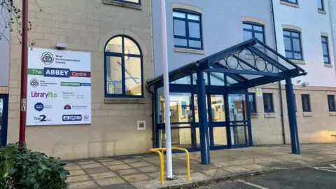 Front entrance to the Abbey Centre - a modern three-storey building in light stone with a blue entrance canopy and glass doors. There is a sign outside showing the logos of various organisations based there. There are paving slabs in front of the building and a bush to the left.