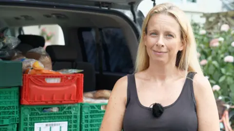 A woman with blonde hair and in a black tank top looking directly at the camera. Behind her is a car boot filled with crates of food.