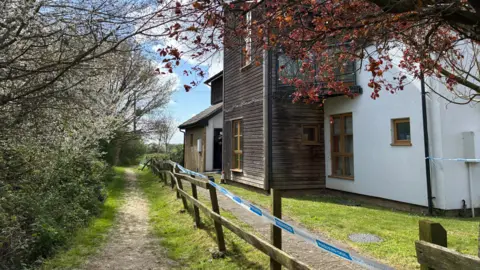 Jenny Kirk/BBC Blue and white police tape runs along the top of a fence. Beyond the fence is a house with white plasterwork and dark wood cladding. To the left, parallel with the fence, is a public footpath, bordered by a line of trees.  