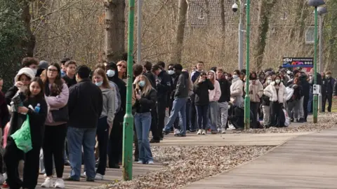PA Media Dozens of young people queue on a concrete path lined with trees. Many are seen wearing face masks.