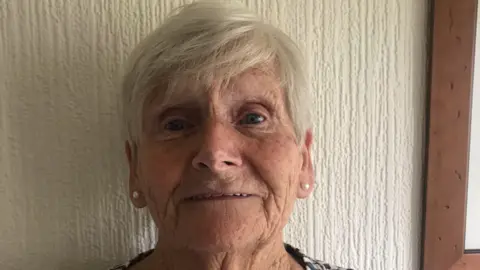 Handout An elderly woman with white short side fringe hair and pearl earrings on against a white wall.