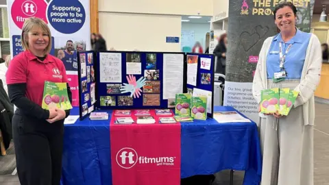 Two women standing holding colourful booklets at either side of a table, covered with a blue tablecloth, with leaflets and posters on it. Heather Gibson is wearing black trousers, a black long-sleeved shirt and a pink shirt. She has shoulder-length blonde hair. Alex Wray is wearing neutral-coloured trousers, a white cardigan and a light blue shirt. She had brown hair tied back. There are posters and logos for Fitmums and Friends.
