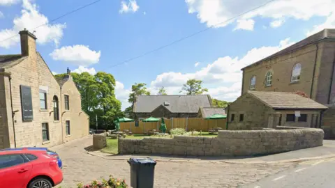 Google A village street scene with traditional stone buildings and a cobbled road curving from left to right. A low, curved stone wall runs along the centre of the image, enclosing a small garden and seating area. A dark wheelie bin stands near the edge of the road in front of the wall and part of a red car is visible on the left.