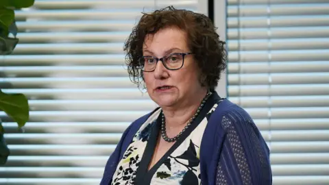 PA Media Dr Hilary Cass, wearing a white and navy dress, blue cardigan and glasses. She has short brown hair. 