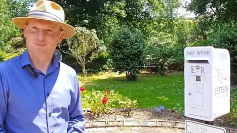 Councillor Loic Rich is wearing a panama hat and blue shirt as he stands near the white post box. It has the logo Post Office and ER on it, with the word Letters down the side. There are red roses and shrubs nearby.