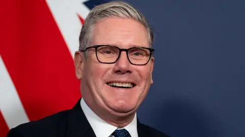 Keir Starmer smiling widely in front of a Union Jack on the wall behind him