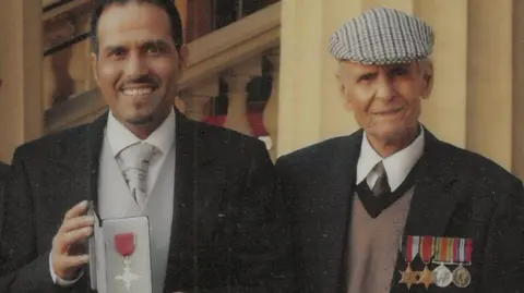 Mashuq Hussain holding his OBE in grey and black tie and tails, standing next to Ghulam Hussain wearing his medals 
