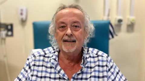 Brian Holmes sits in a blue cushioned chair in a hospital. His mouth is slightly open. He has wavy grey hair and has a grey beard around his chin and mouth. He is wearing a white, blue and black gingham-style shirt.