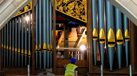 Huge organ pipes at the top of the organ. There's a man in a high-visibility jacket and construction hat on a ladder.