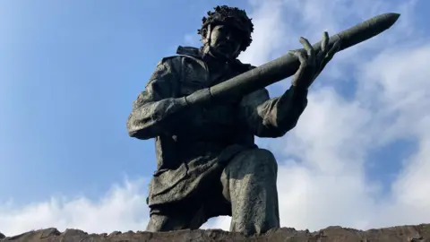 A bronze statue of a solider holding a weapon, bathed in sunlight and against a blue sky 